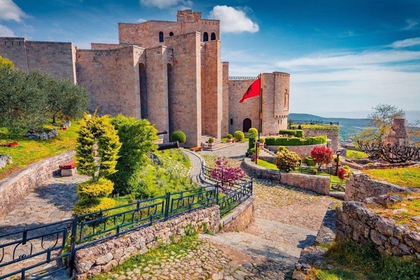 A historic stone castle with a red flag, surrounded by well-maintained gardens and a stone pathway. 1