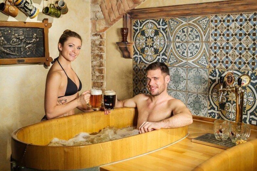 Two people in a wooden bathtub with beer mugs, surrounded by tiled walls and a beer tap. 5
