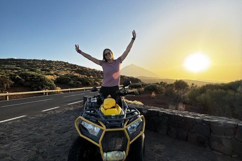 A person on a yellow quad bike with arms raised, a road, and a mountainous landscape in the background. 1