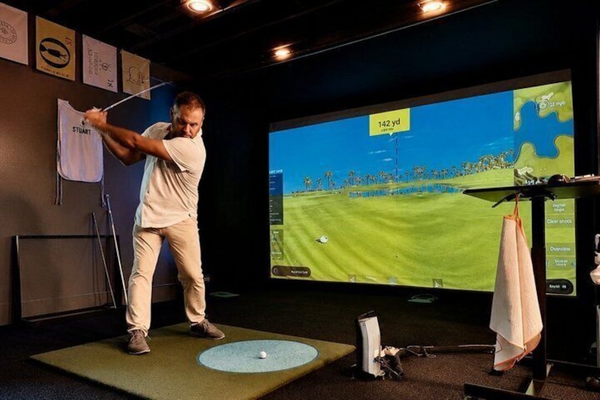 A person practicing golf swings on a mat with a large screen displaying a golf course in the background. 5