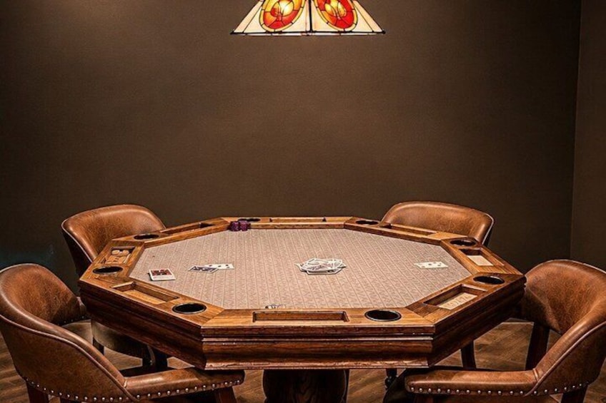 A poker table with cards and chips, surrounded by leather chairs, in a dimly lit room with a stained glass window. 3