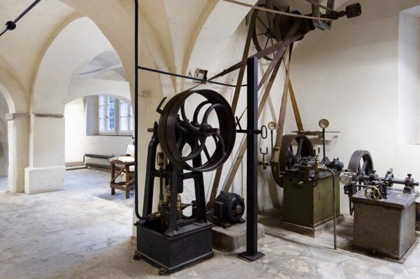 An industrial setting with machinery, including a large pulley system and smaller devices, in a room with arched ceilings and a stone floor. 5