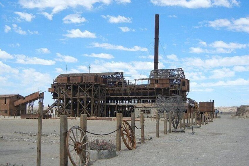 An abandoned industrial structure with a tall chimney, surrounded by wooden posts and a chain-link fence. 1