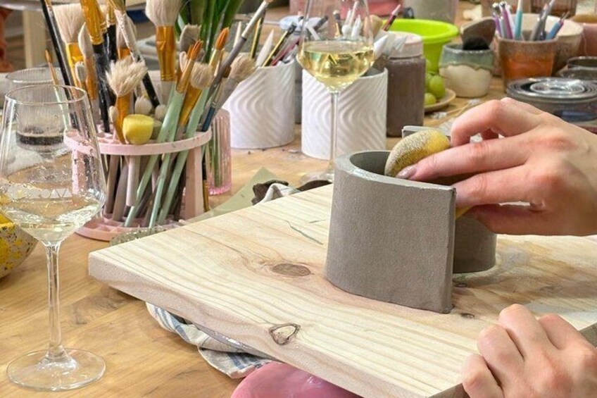 A person holding a brush, a cup, and a glass on a wooden table with art supplies. 4