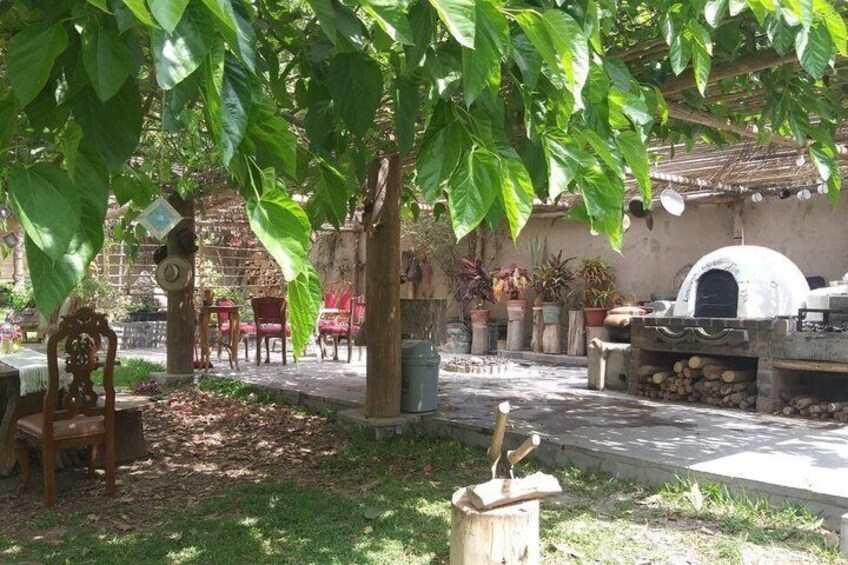 A rustic outdoor setting with a wooden table, chairs, a tree, a brick oven, and potted plants. 5