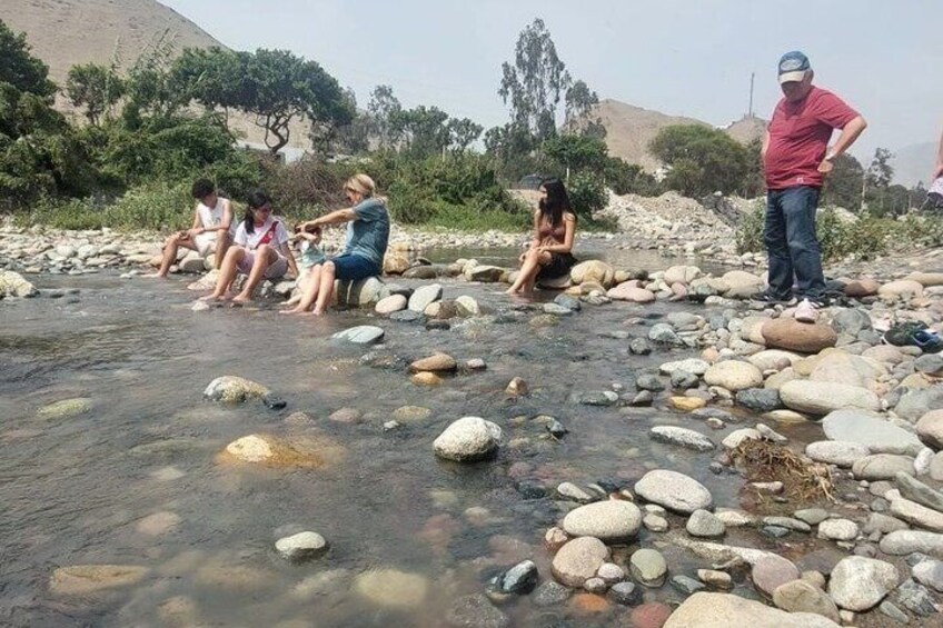People sitting by a clear stream with rocks, surrounded by trees and hills. 2