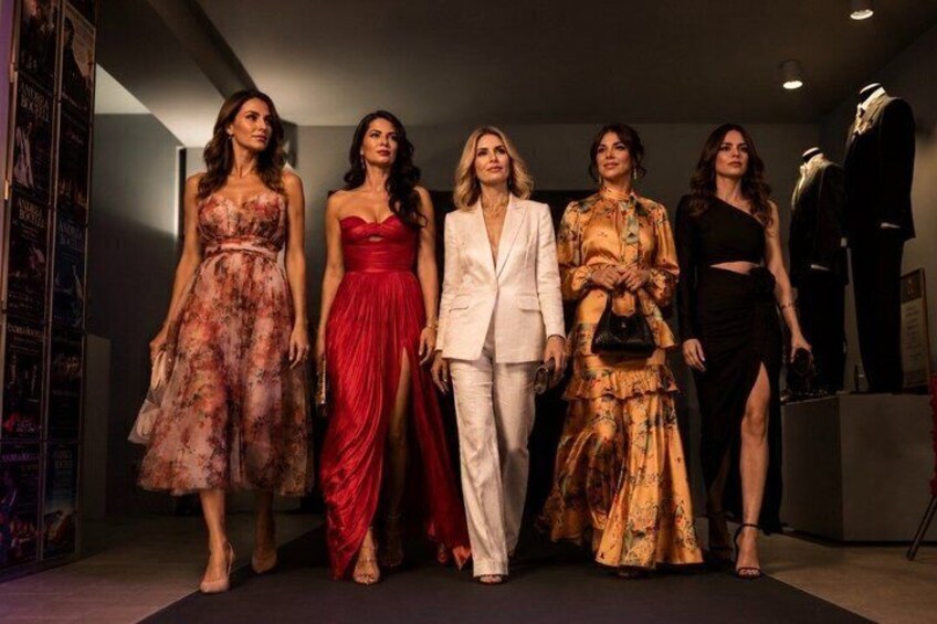 A group of women in formal attire standing in a room with a mannequin and posters on the wall. 2