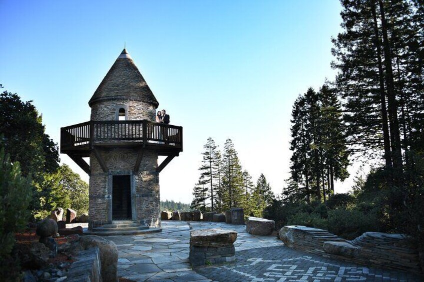 A stone tower with a balcony, surrounded by a stone pathway and tall trees. 3