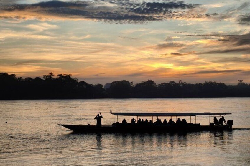 A boat with passengers at sunset, calm water, and a silhouette of trees on the horizon. 1