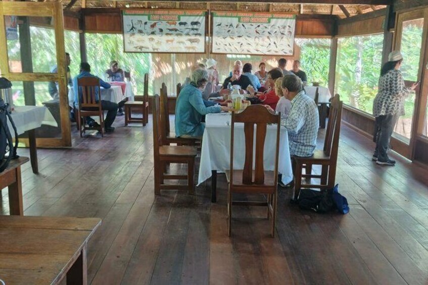 A group of people dining at wooden tables in a covered outdoor area with a bird identification chart on the wall. 3