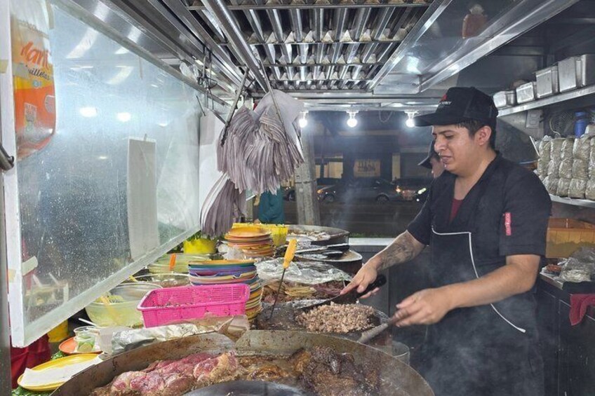 A person cooking meat in a large pan at a food stall. 4