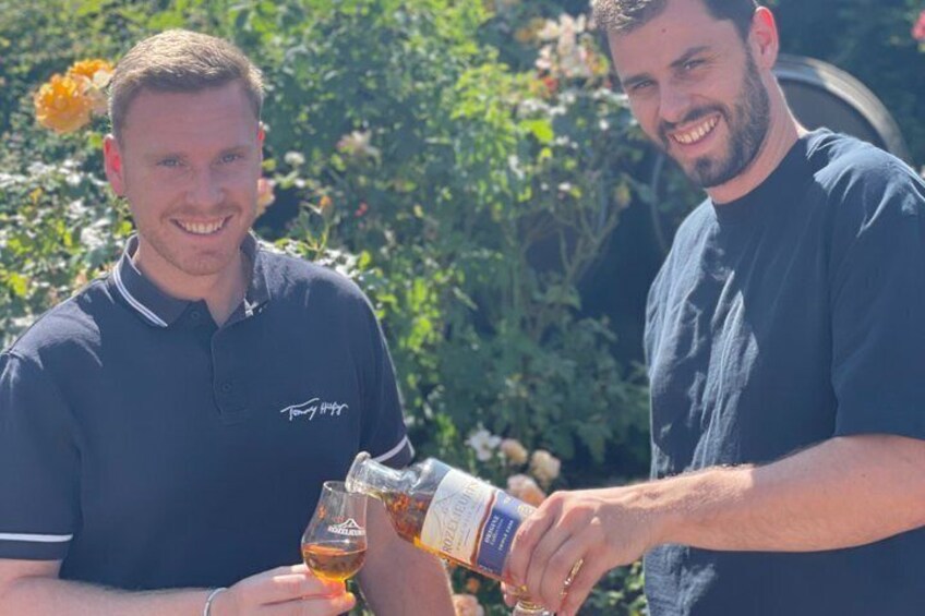 Two individuals outdoors, one pouring a beverage into a glass, with greenery and flowers in the background. 3
