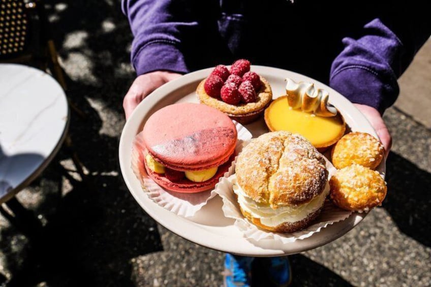 A person holding a tray with macarons, a tart, a cheesecake, and a pastry. 1