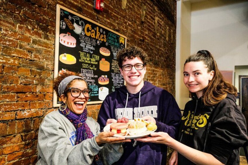 Three people holding a plate of food in front of a chalkboard menu with various cake options. 4