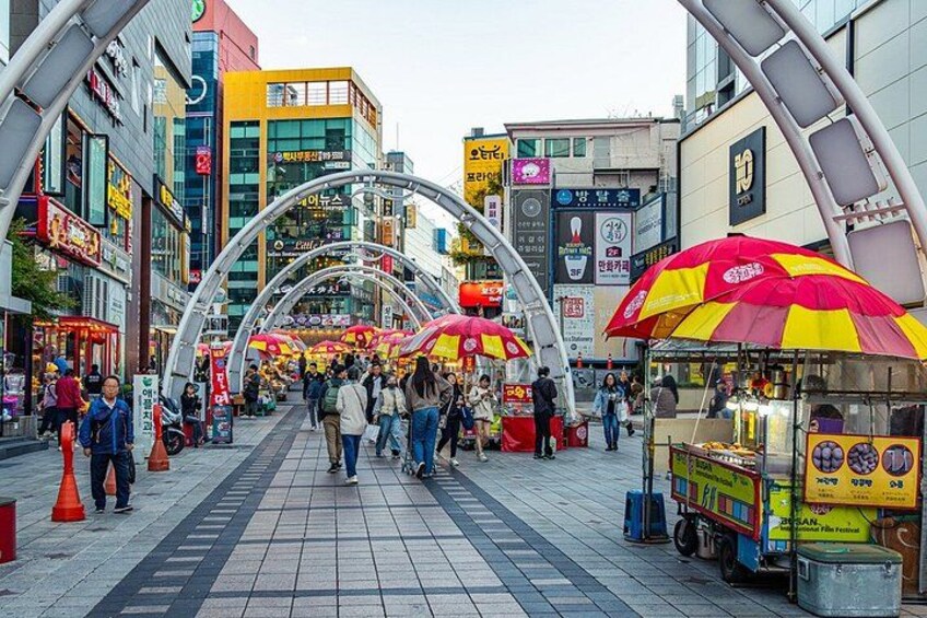 A busy urban street with pedestrians, food stalls, and modern buildings. 5