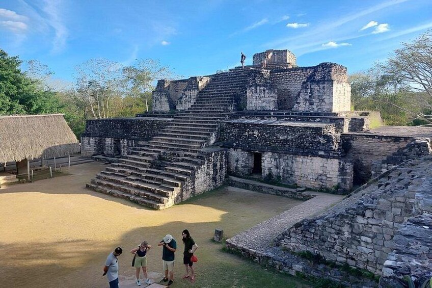 An ancient Mayan pyramid with a staircase leading to the top, surrounded by a sandy area and trees. 1
