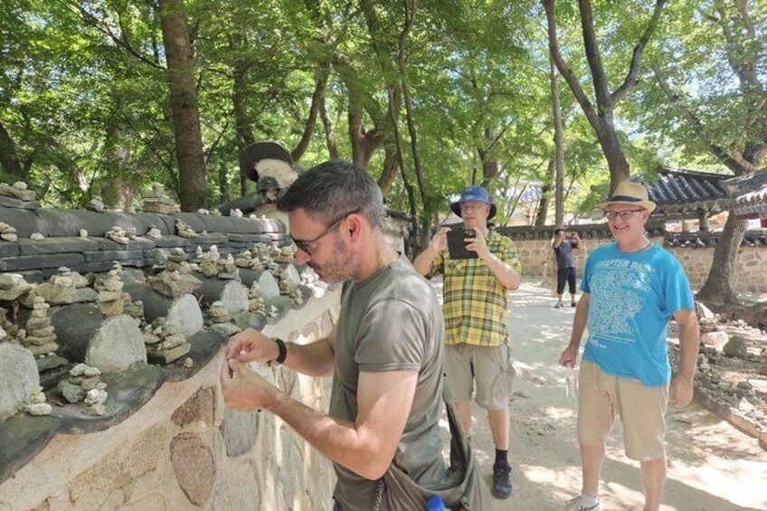 A man examining rocks, a person taking a photo, and a traditional structure in a wooded area. 2