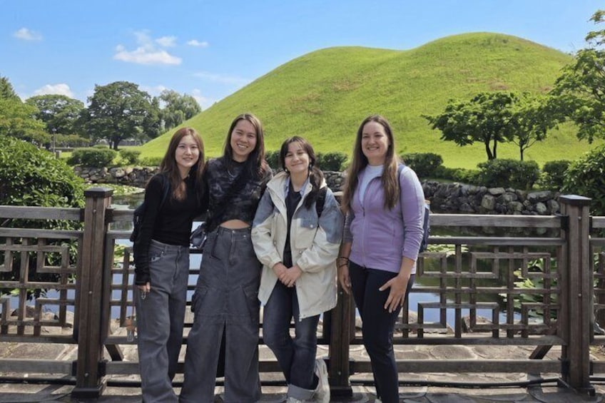 Four people standing in front of a wooden fence with a green hill and trees in the background. 1
