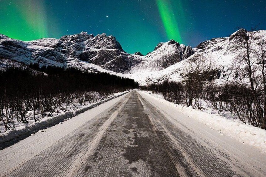 A snow-covered road flanked by mountains under a starry sky with the aurora borealis. 5