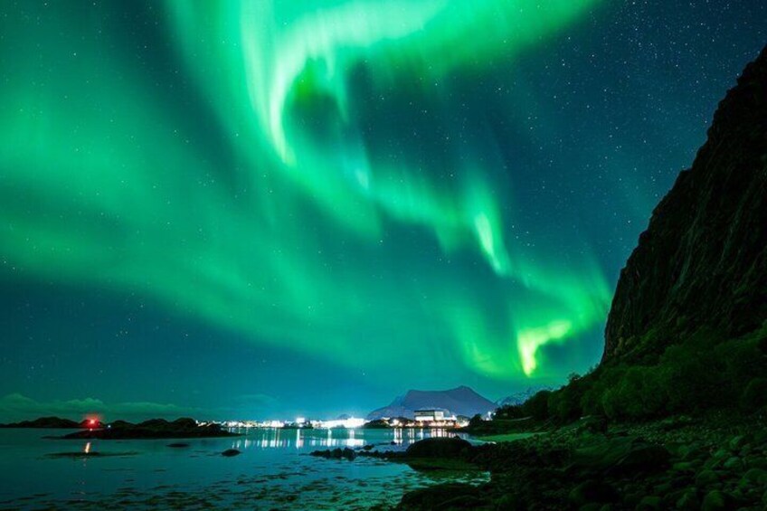 The Northern Lights over a coastal landscape with a mountain and a lit building in the distance. 1