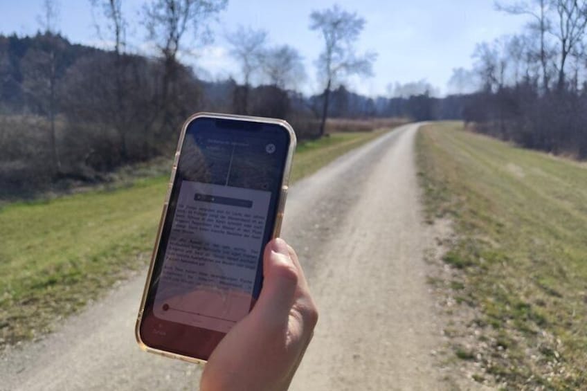 A hand holding a smartphone with a map displayed, standing on a rural dirt road with trees on either side. 3
