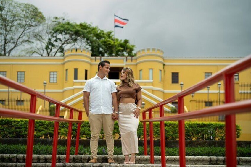 A couple standing on a staircase with a yellow building and a flag in the background. 1