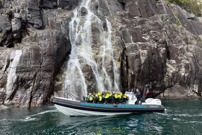A boat with people wearing life jackets, a waterfall, and rocky cliffs in the background. 1