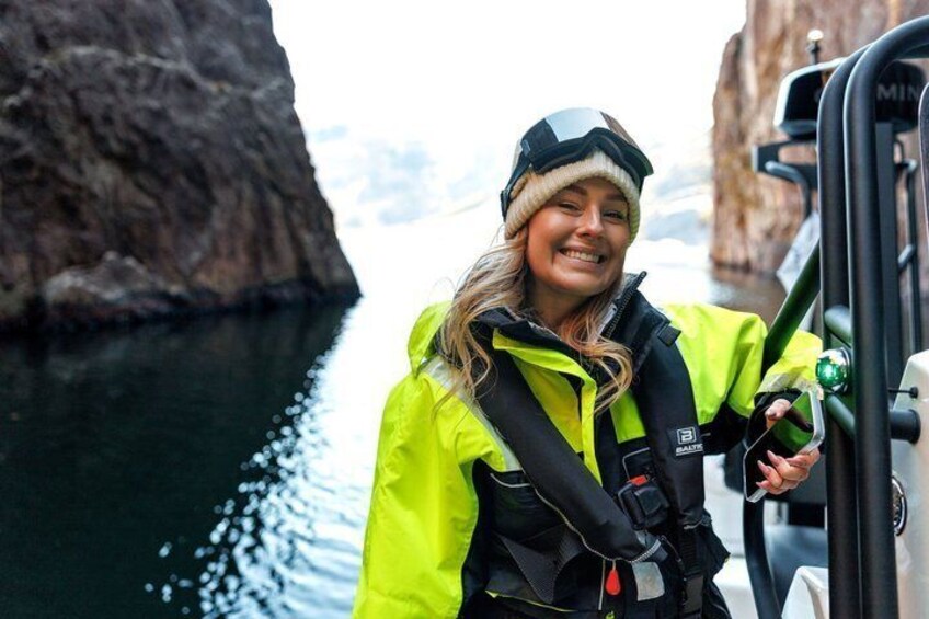 A person in a bright yellow jacket and helmet, standing by a boat with a rocky shoreline in the background. 2