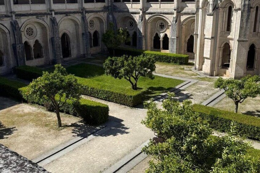 A courtyard with a central pathway, surrounded by neatly trimmed hedges and trees, with a historic building featuring arched walkways in the background. 5