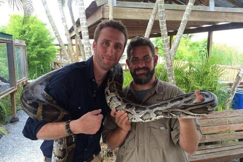 Two people holding a large snake in an outdoor setting with wooden structures and greenery. 1
