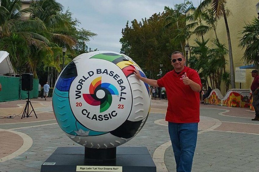 A man in a red shirt standing next to a large, colorful baseball-themed sculpture with the text 'WORLD BASEBALL CLASSIC' and numbers '20' and '23'. 4