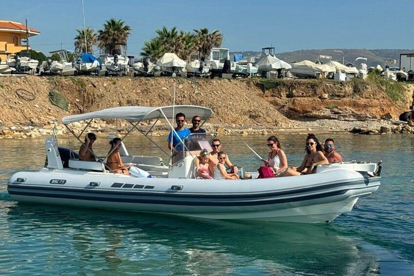 A small motorboat with people on board, a shoreline with parked boats, and a clear blue sky. 1