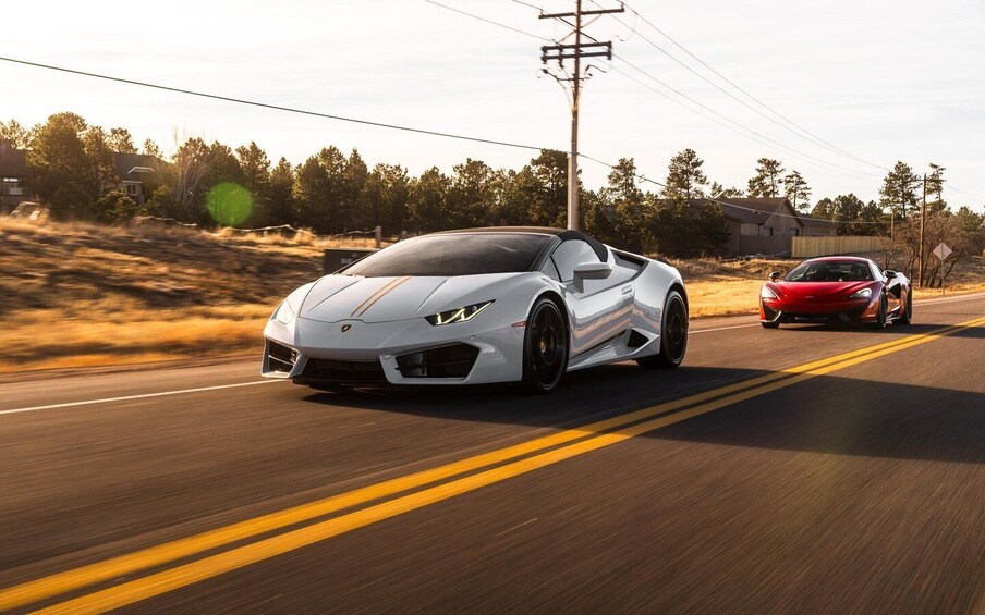 Two sports cars driving on a road with a double yellow line, surrounded by trees and a wooden structure. 2