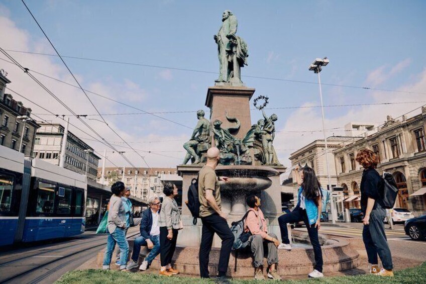 A public square with a statue fountain, tram tracks, and urban buildings. 1