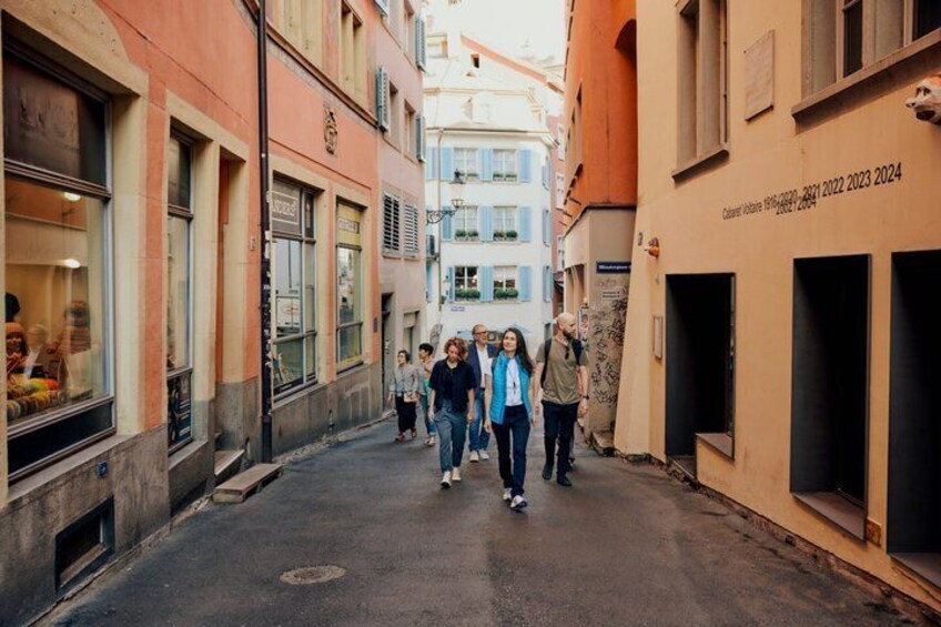 A narrow street with buildings on either side, a group of people walking, and a sign with a date range. 3