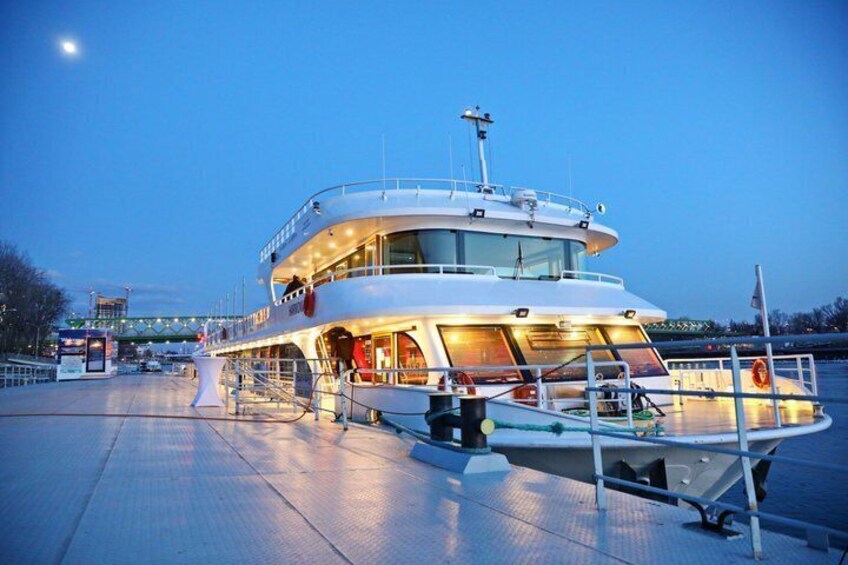 A large, multi-deck boat docked at a pier with a city skyline in the background. 1