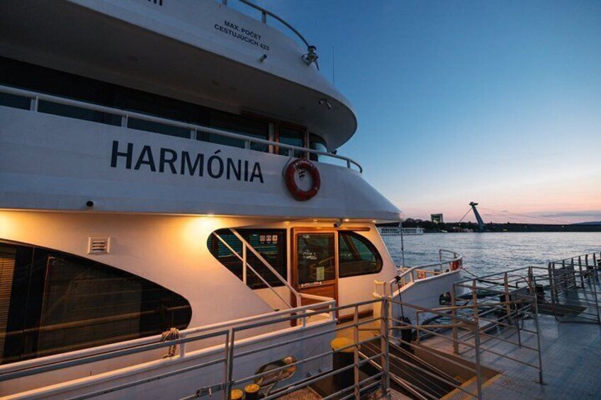 A large white boat named 'HARMÓNIA' docked at a pier with a city skyline in the background. 3