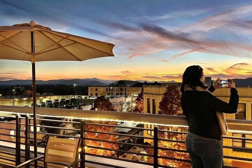 A person taking a photo of a sunset over a cityscape with buildings and a river. 1