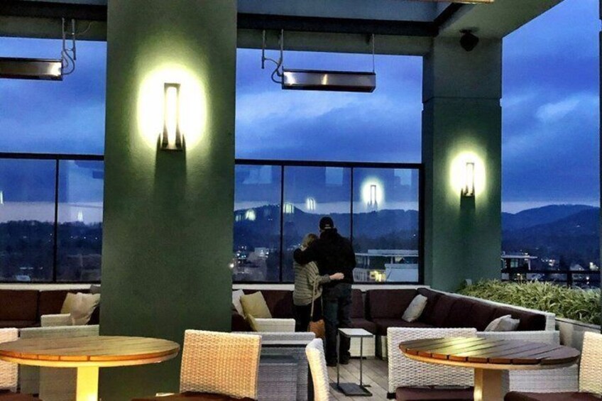 An outdoor seating area with round tables and chairs, large windows, and a view of a cityscape at dusk. 2