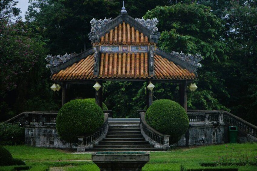 A traditional pavilion with a tiled roof, surrounded by a stone staircase and two spherical topiary bushes. 5