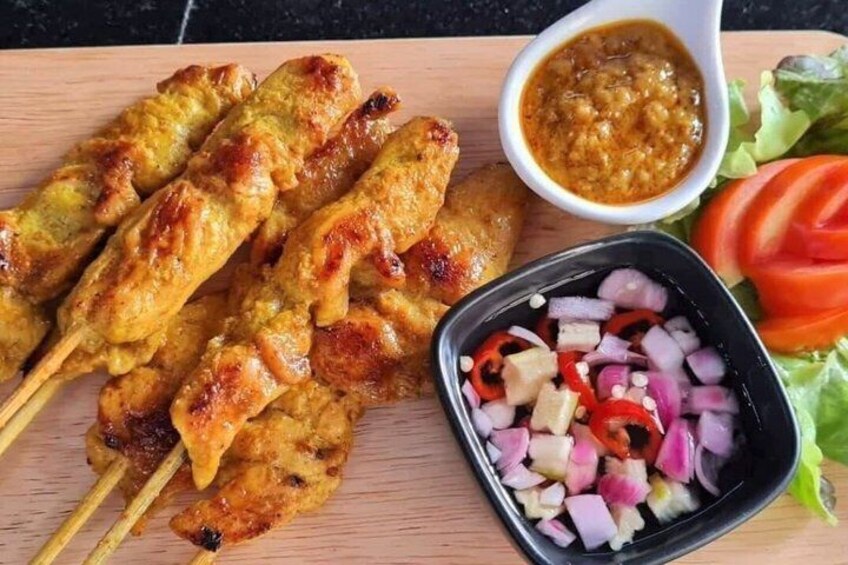 Grilled chicken skewers, a bowl of sauce, diced onions, and sliced tomatoes on a wooden board. 3