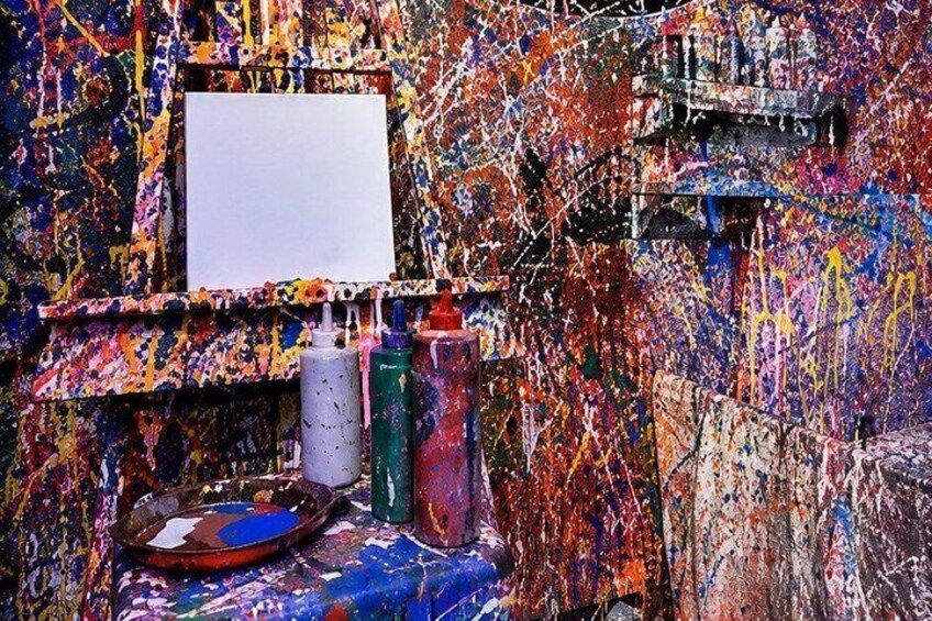 A chaotic art studio with a white canvas, paint bottles, and a palette. 3