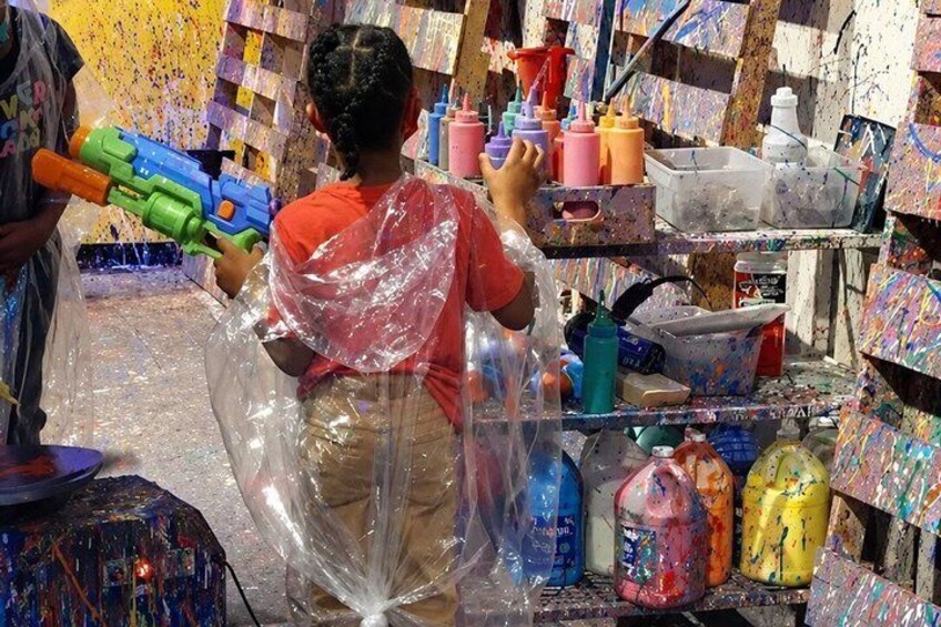 A person in a red shirt and plastic wrap, surrounded by paint bottles and containers in a room with a colorful, paint-splattered wall. 4