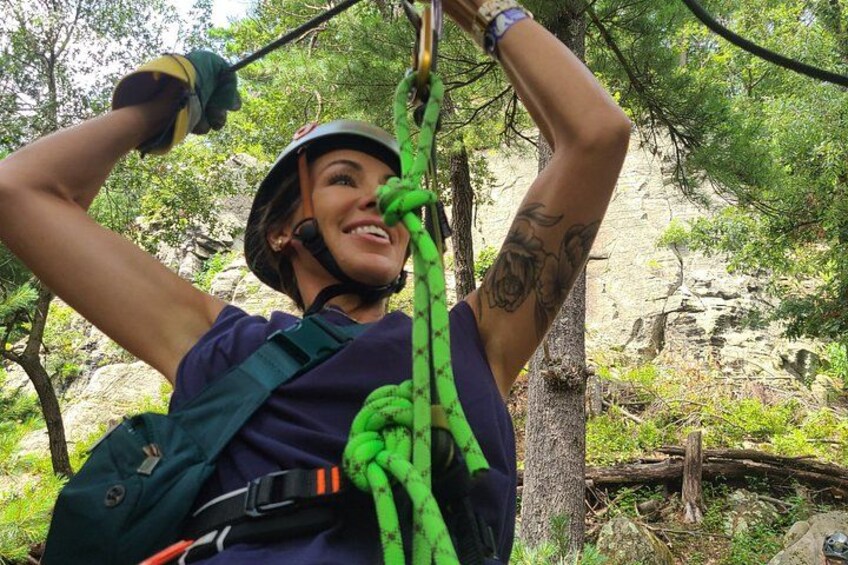 A person rappelling down a tree, wearing a helmet and harness, surrounded by greenery and rocks. 1