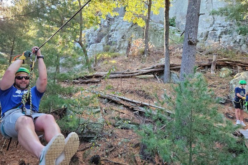 A person sitting on a rope swing in a forested area with trees and a rocky background. 3