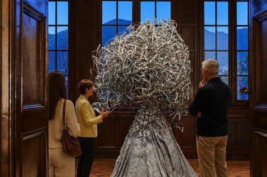 A group of people observing a large, abstract sculpture in a room with wooden paneled walls and large windows. 1