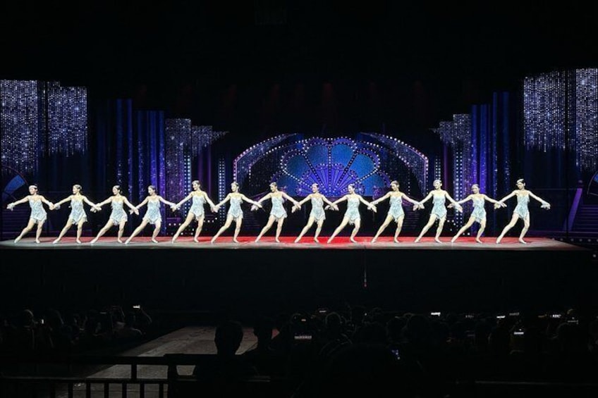 A group of dancers in light blue costumes performing on a stage with a large, illuminated backdrop. 3