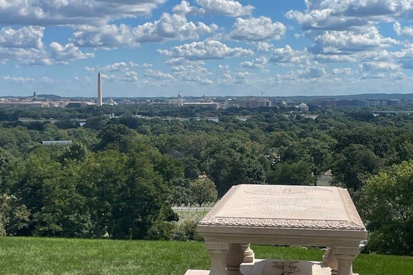 A stone monument in a grassy area with a view of the Washington Monument and surrounding trees. 5
