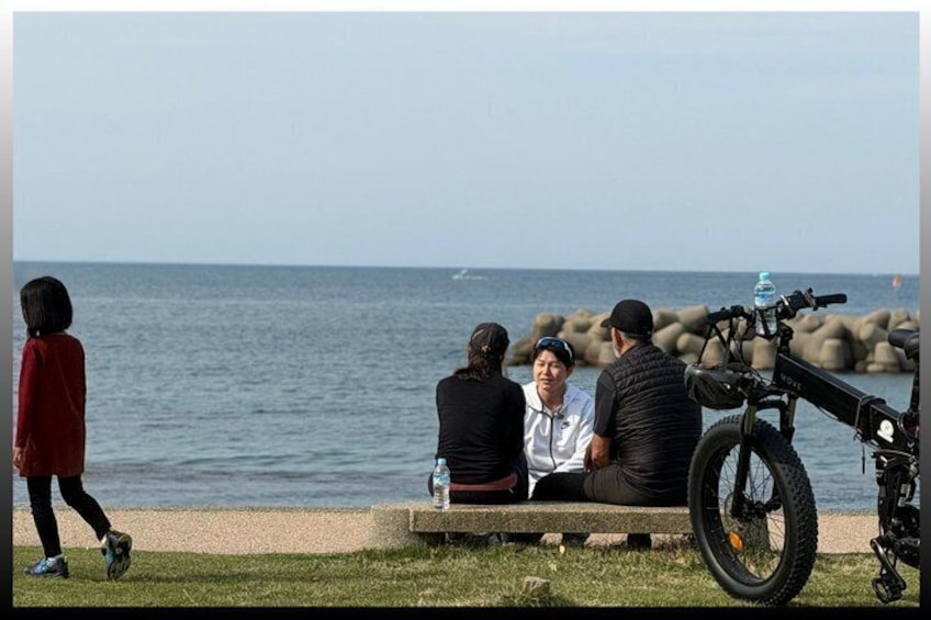 A group of people sitting on a bench by the sea, a motorcycle parked nearby, and a person walking in the foreground. 2
