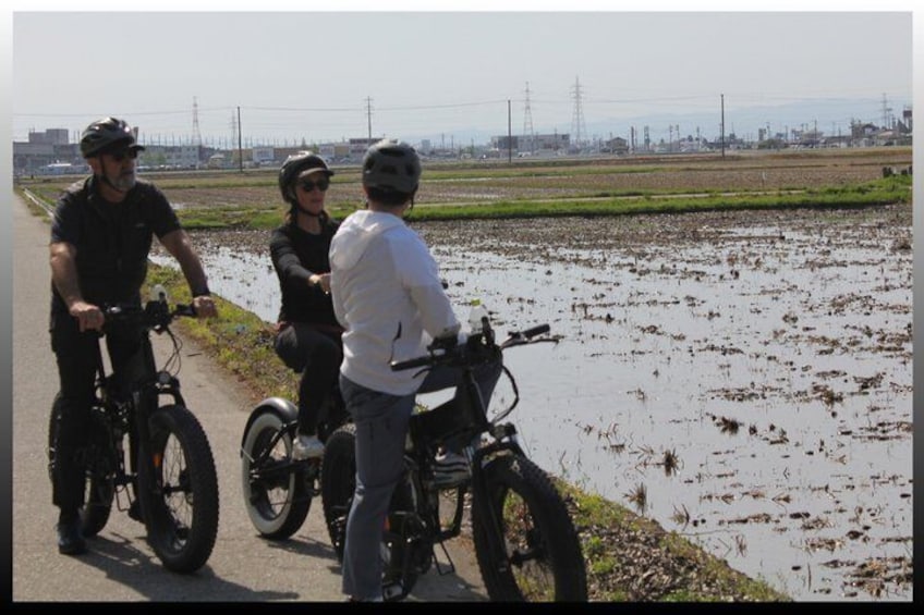 Three people on bicycles near a flooded field with a distant industrial area. 4